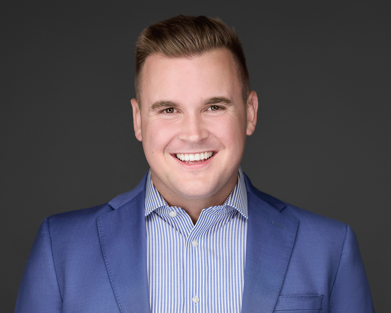 Professional headshot of Jayden Bell in blue suit – Dental Coach USA Jayden Bell, Co-Founder and CEO of Dental Coach USA, smiling in a professional headshot while wearing a blue suit jacket and striped shirt against a dark background – Dental Coach USA