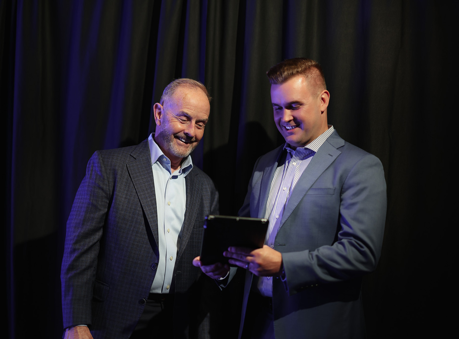 Bob Affleck and Jayden Bell reviewing information on tablet – Dental Coach USA Bob Affleck, Co-Founder and CSO, and Jayden Bell, Co-Founder and CEO of Dental Coach USA, smiling as they review information on a tablet while standing in front of a dark curtain backdrop – Dental Coach USA